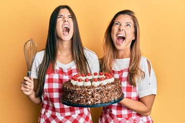 Sticker - Hispanic family of mother and daughter wearing baker apron holding homemade cake angry and mad screaming frustrated and furious, shouting with anger looking up.