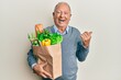 Senior caucasian man holding paper bag with bread and groceries pointing thumb up to the side smiling happy with open mouth