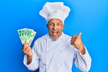 Poster - Middle age grey-haired man wearing professional cook uniform holding israel shekels banknotes pointing thumb up to the side smiling happy with open mouth