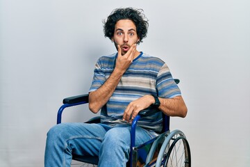 Poster - Handsome hispanic man sitting on wheelchair looking fascinated with disbelief, surprise and amazed expression with hands on chin