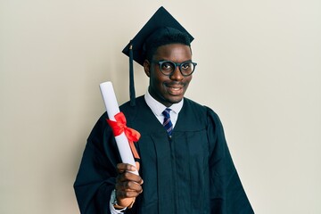 Wall Mural - Handsome black man wearing graduation cap and ceremony robe holding diploma looking positive and happy standing and smiling with a confident smile showing teeth