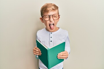 Little caucasian boy kid reading a book wearing glasses sticking tongue out happy with funny expression.