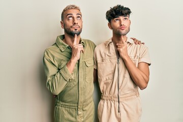 Poster - Homosexual gay couple standing together wearing casual jumpsuit thinking concentrated about doubt with finger on chin and looking up wondering