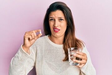 Wall Mural - Beautiful hispanic woman holding pill and glass of water in shock face, looking skeptical and sarcastic, surprised with open mouth