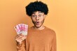 © Krakenimages.com - Young african american man with afro hair holding 100000 indonesian rupiah scared and amazed with open mouth for surprise, disbelief face