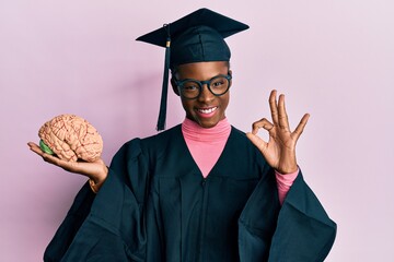 Sticker - Young african american girl wearing graduation cap and ceremony robe holding brain doing ok sign with fingers, smiling friendly gesturing excellent symbol