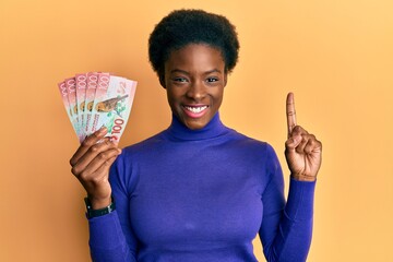 Canvas Print - Young african american girl holding 100 new zealand dollars banknote smiling with an idea or question pointing finger with happy face, number one