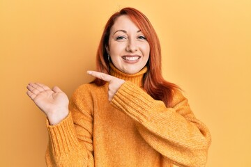 Poster - Beautiful redhead woman wearing casual winter sweater over yellow background amazed and smiling to the camera while presenting with hand and pointing with finger.