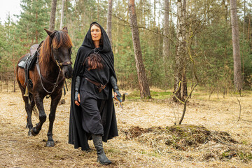 Wall Mural - beautiful black dressed woman in historical costume with bow and arrow walks through the woods with her horse