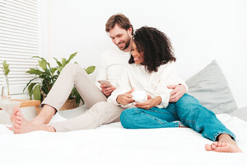 Smiling beautiful woman and her handsome boyfriend. Happy cheerful multiracial family. Multiethnic models lying in bed and hugging in white interior.Looking at cellphone screen. Using smartphone apps