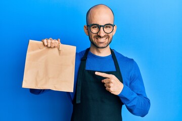 Wall Mural - Young hispanic man wearing waiter uniform holding take away paper bag smiling happy pointing with hand and finger