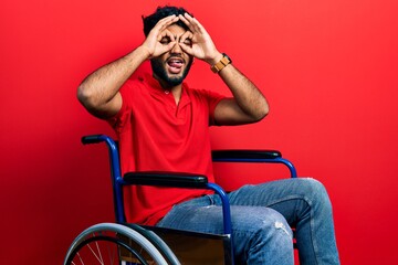 Canvas Print - Arab man with beard sitting on wheelchair doing ok gesture like binoculars sticking tongue out, eyes looking through fingers. crazy expression.