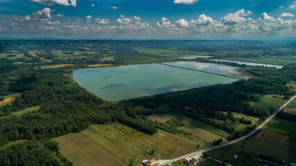 Wall Mural - Aerial view of a road amid agricultural fields with a blue lake and buildings under a cloudy sky