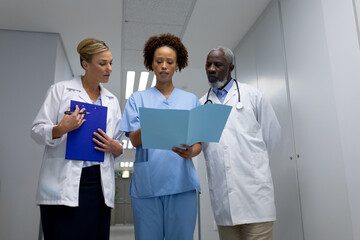 Wall Mural - Three diverse male and female doctors standing in hospital corridor looking at medical documentation