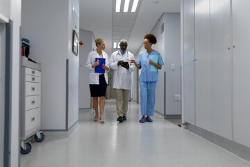 Wall Mural - Three diverse male and female doctors walking through hospital corridor looking at digital tablet