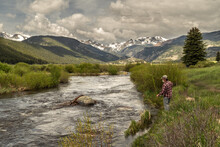 A Person, Mature, Caucasian, Fly Fishing On A River With View Of Snow Covered Mountains And Dramatic Cloudy Sky For Background, Big Thompson River, Rocky Mountain National Park, Colorado