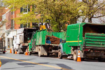 A convoy of trucks operated by a local landscaping company are parked on the street by a park at and urban location. They are loaded with piles of manure, fertilizer and compost for spring garden care