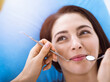 © rogerphoto - Smiling brunette woman being examined by dentist at sunny dental clinic. Hands of a doctor holding dental instruments near patient's mouth. Healthy teeth and medicine concept