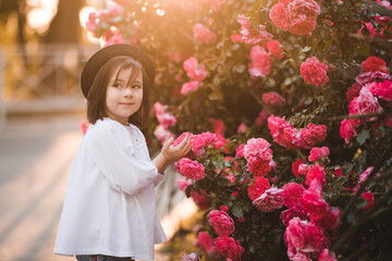 Wall Mural - Cute stylish child girl 5-6 year old wear trendy clothes and hat posing over flower rose outdoors in park. Having fun. Childhood. Happiness.