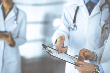 Unknown doctors, two men and a woman, discuss medical exam resoults, while standing at hospital office. Physicians using clipboards for filling up medication history records. Perfect medical service