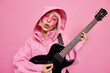 © WHstudio Leushin N - Carefree female singer enjoys music sings favorite song plays electrical guitar dressed in anorak feels glad poses against pink background takes part in festival. Rock star demonstrates her talent