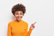 Beautiful curly haired Afro American woman suggests to clickon link points on blank copy space shows way stands over white background wears basic orange jumper. People and advertising concept