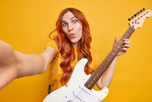 Funny Redhead Woman Makes Surprised Grimace At Camera Makes Selfie Portrait While Playing Acoustic Guitar Wears Casual T Shirt And Hat Isolated Over Vivid Yellow Background. Lifestyle And Hobby
