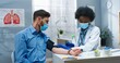 © VAKSMANV - Portrait of African American female pretty doctor in medical mask measuring blood pressure of Caucasian young handsome male patient in cabinet in clinic. Coronavirus pandemic concept