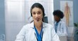 © VAKSMANV - Close up portrait of Caucasian pretty young woman doctor in white medical coat waving hand looking at camera and speaking in headset sitting in hospital cabinet. Healthcare, video consultation