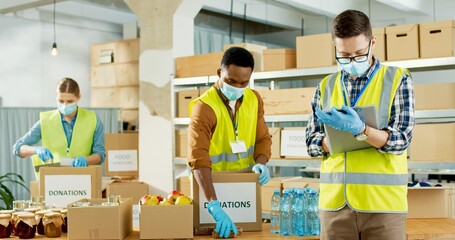 Wall Mural - Portrait of young Caucasian male manager in medical mask writing checking donation list. Mixed-race volunteer workers working in shipping delivery charitable stock organization packing donations box