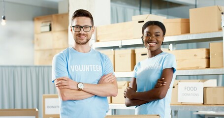 Wall Mural - Portrait of happy cheerful young Caucasian handsome guy and beautiful African American woman volunteers standing in charity center stock looking at camera and smiling. Volunteering activity concept