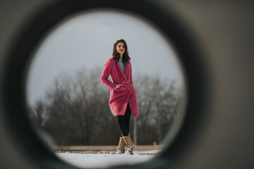 Sticker - Full body shot of a beautiful cute woman wearing a pink coat posing in a snowy park through a hole