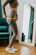 © shevtsovy - slim Woman waist Measuring With Tape at home. Beautiful woman checking success of weight loss program and measuring body in living room. slimming and weight loss concept.