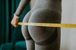 © shevtsovy - Fitness Woman Measuring butt With Tape at home. strong buttocks concept. Beautiful woman checking success of weight loss program and measuring butt and hips in front of mirror in living room.