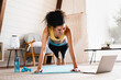 © InsideCreativeHouse - Fit sporty african-american young woman standing in plank position using laptop at home. Cheerful African woman stretching up In front of laptop, doing home workout,