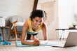 © InsideCreativeHouse - Slim strong african-american woman standing in plank position looking at the laptop indoors. African female warming up for domestic fitness training, exercising on fitness mat in living room