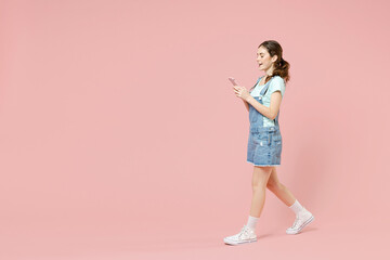 Wall Mural - Full length side view young smiling caucasian woman 20s wear trendy denim clothes blue t-shirt hold mobile cell phone walk isolated on pastel pink background studio portrait. People lifestyle concept