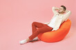 © ViDi Studio - Full length young smiling happy overjoyed joyful fashionable man 20s in jacket white t-shirt sitting in bean bag chair resting hold hand behind neck isolated on pastel pink background studio portrait
