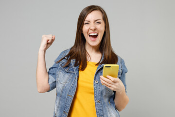 Wall Mural - Young fun smiling happy caucasian woman 20s in casual trendy denim jacket yellow t-shirt using mobile cell phone do winner gesture clench fist chat online isolated on grey background studio portrait