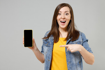 Poster - Young excited surprised caucasian woman in casual trendy denim jacket yellow t-shirt using point index finger on mobile cell phone with blank screen workspace area isolated on grey background studio