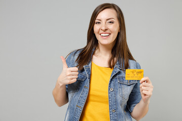 Wall Mural - Young fun smiling happy caucasian woman 20s in casual trendy denim jacket yellow t-shirt holding in hand credit bank card look camera show thumb up gesture isolated on grey background studio portrait