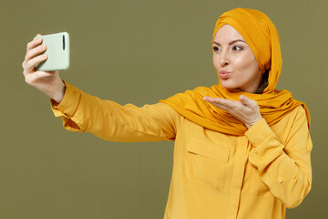 Poster - Young girlfriend arabian asian muslim woman 20s wear abaya hijab yellow clothes do selfie shot on mobile phone blowing air kiss isolated on olive green background People uae islam religious concept