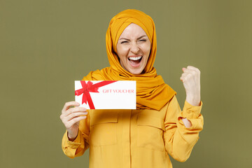 Sticker - Young arabian asian muslim woman in abaya hijab yellow clothes hold gift voucher flyer mock up do winner gesture isolated on olive green background studio portrait People uae islam religious concept.