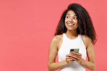Young smiling happy dreamful thoughtful wistful pensive african american woman 20s wear casual white tank shirt hold in hand using mobile cell phone chat online look aside isolated on pink background