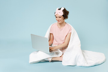 Young woman in pajamas jam sleep eye mask rest at home sit wrap cover under blanket duvet use laptop pc computer chat social network isolated on pastel blue background Good mood night bedtime concept.