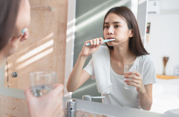 Young Asian woman brushing teeth and looking in the mirror, holding water glass, towel on the shoulder on bathroom, Concept oral hygiene and health care.