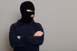 A criminal man in bandit mask and a black hoodie stands with folded hands on his chest, looking to the side, copy space for advertising text, isolated on gray background.