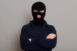 Self-confident criminal male posing isolated on a gray background, wearing a black hoodie and a bandit mask, looking at the camera, holding his hands folded on his chest.