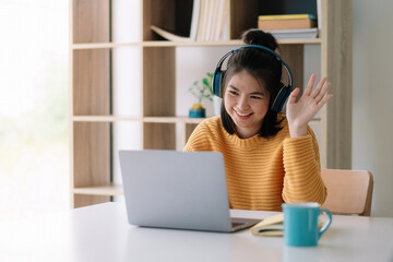 happy young asian ethnicity female manager wearing wireless headphones, looking at laptop screen, holding pleasant conversation with partners clients online, working remotely at workplace.