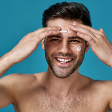 Happy Brunette Guy Smiling At Camera While Applying White Cream On His Face, Posing Isolated Over Blue Background
